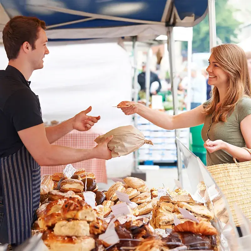 Een verkoper overhandigt een bruin papieren zak aan een klant bij een marktstand met verschillende gebakken goederen.
