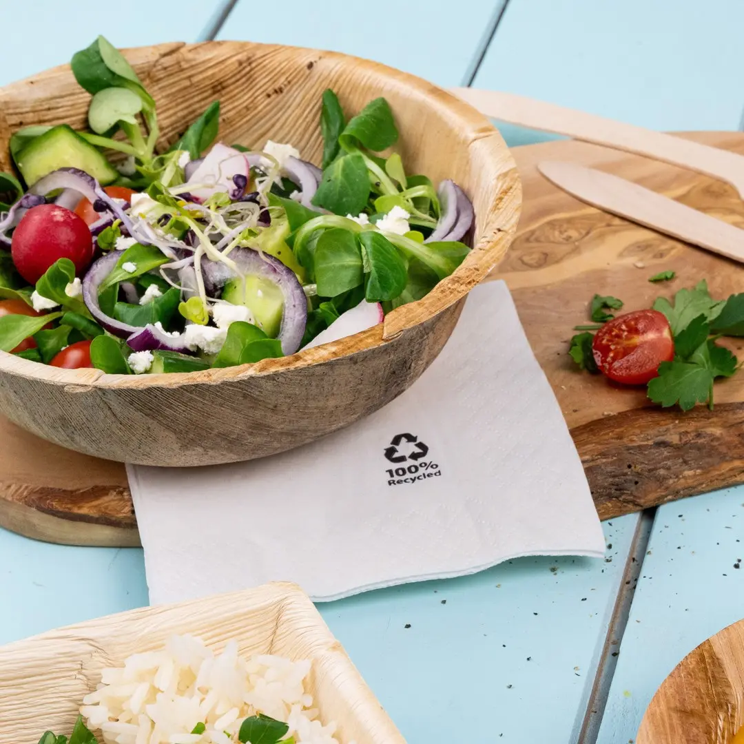 Salade in een houten kom met een servet met het label "100% Recycled", naast rijst en een cherrytomaat op een houten plank.