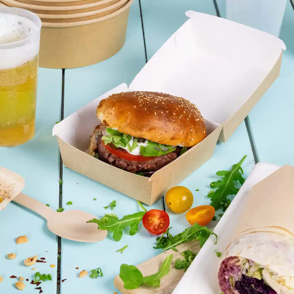 Een hamburger met sesambroodje, sla, tomaat en saus in een bruine afhaaldoos, vergezeld van groenten en bier.