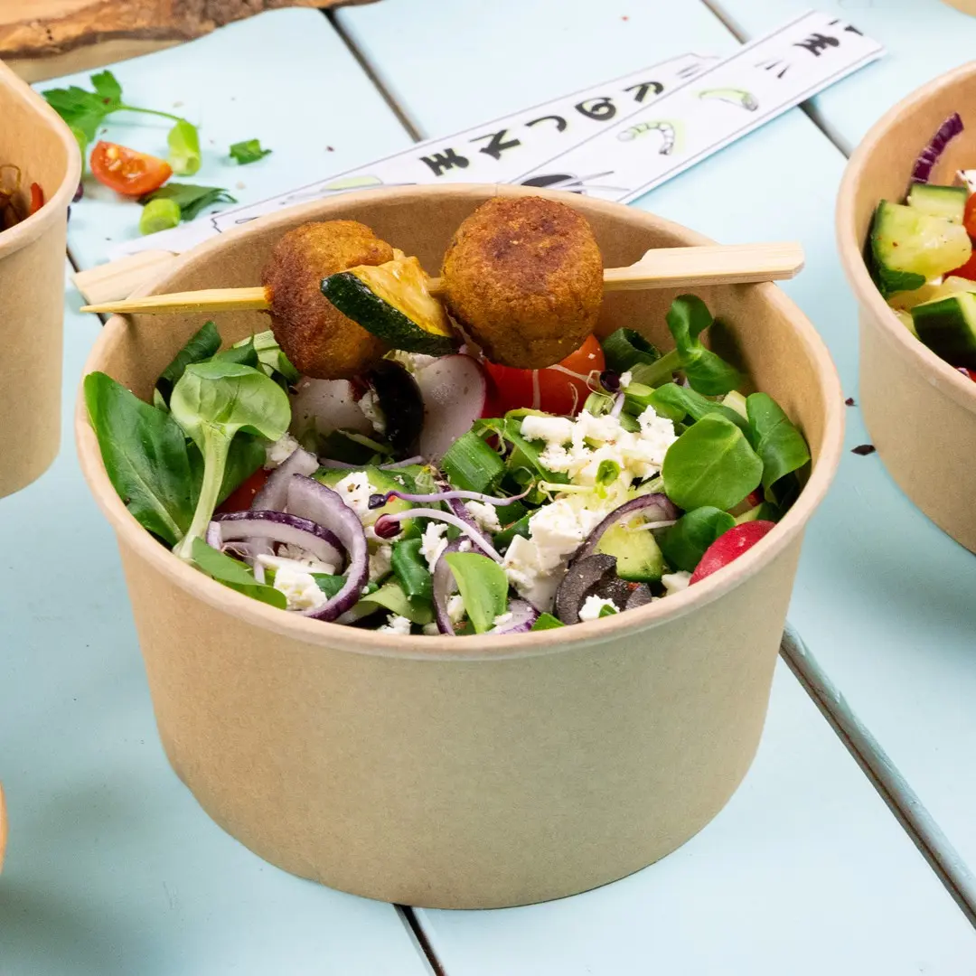 Salade in een bruine papieren kom met groenten, tomaten, uien, olijven, feta en twee falafelballen op een spies.