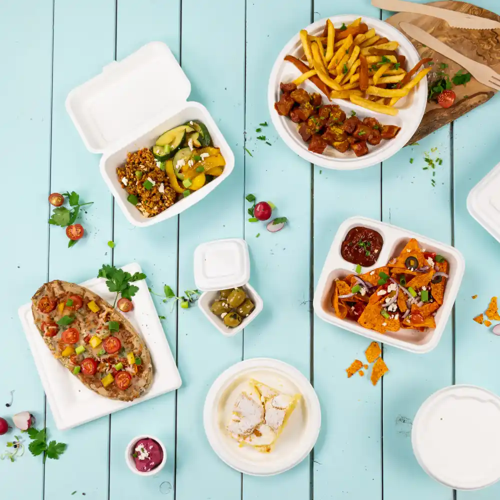 Assortiment wegwerp voedselcontainers met verschillende gerechten, waaronder frietjes, nacho's, salade en dessert op een blauwe houten tafel.