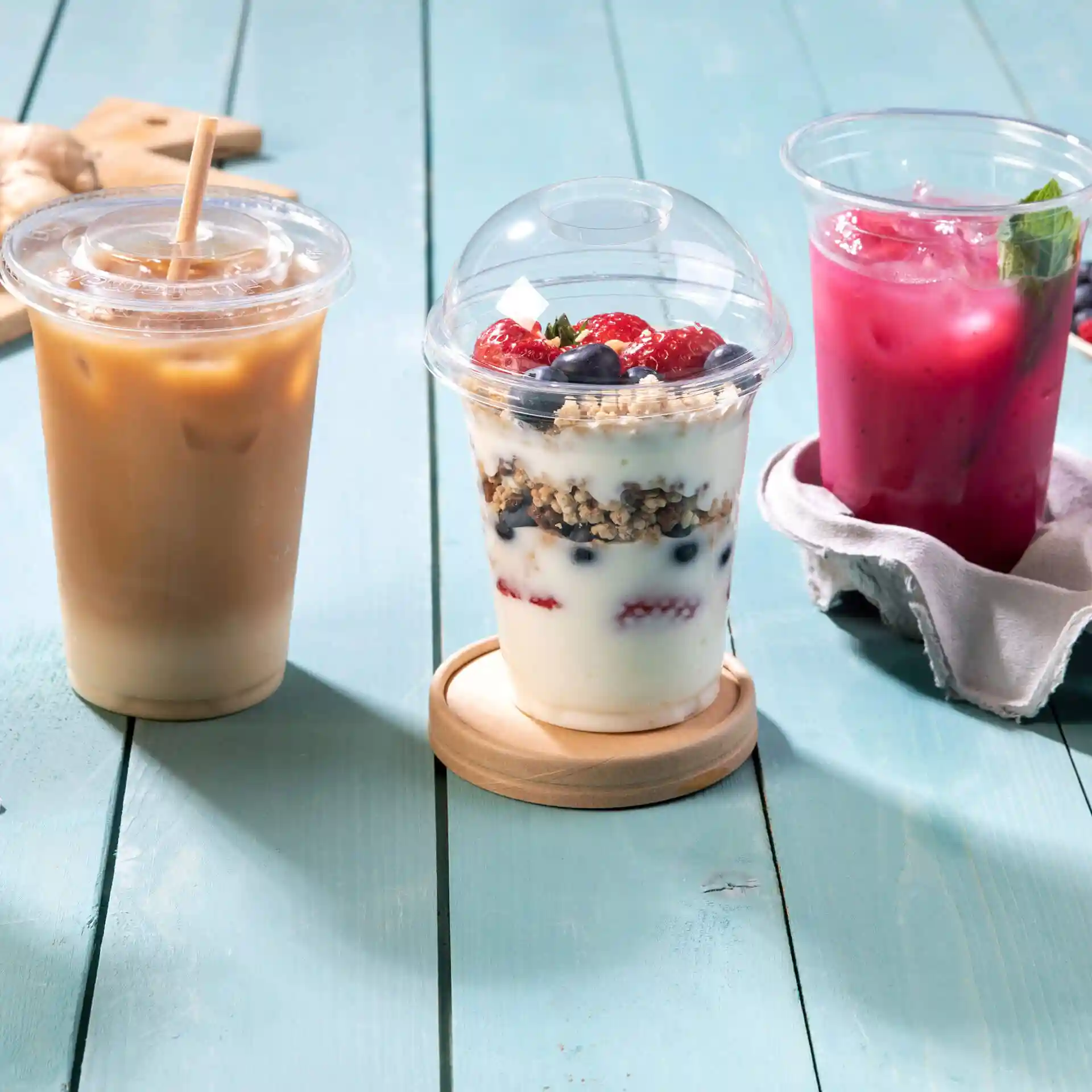Afbeelding van drie dranken: een iced coffee, een yoghurtparfait met bessen en een roze drankje, allemaal in transparante bekers op een blauwe houten ondergrond.