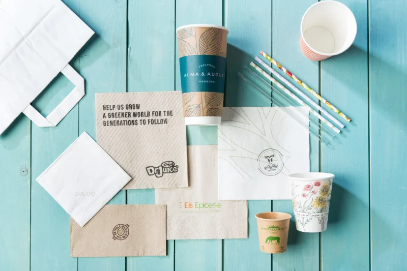 Diverse verpakkingsartikelen zoals papieren bekers, servetten en rietjes op een turkooise houten oppervlak.