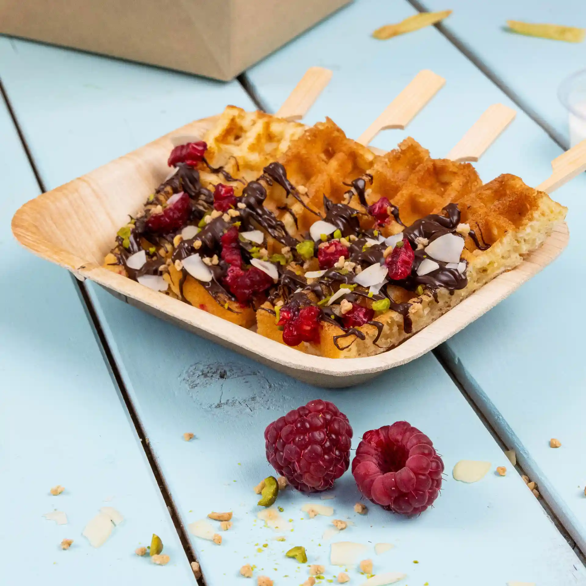 Diepe rechthoekige palmbladplaat met wafels, getopt met chocolade, frambozen en noten, op een blauwe houten ondergrond.