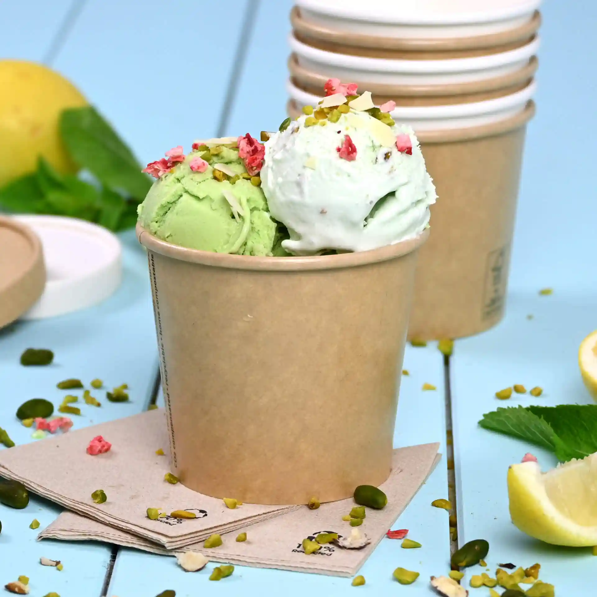Bruine papieren ijsbeker gevuld met groen en wit ijs, getopt met noten en roze sprinkles, op een blauwe ondergrond.