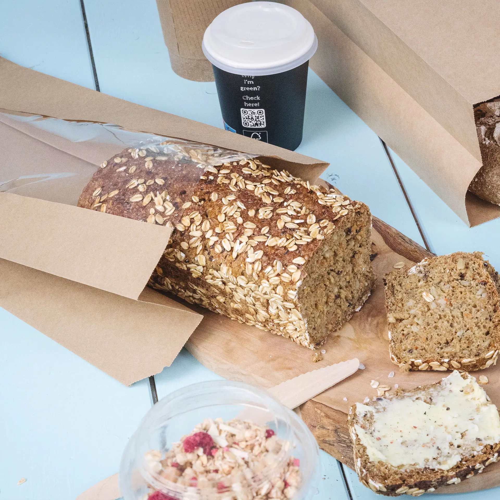 Een brood van bruin brood met havermouttopping, gedeeltelijk gesneden, naast een drinkbeker en een container met granola, alles op een blauwe ondergrond.