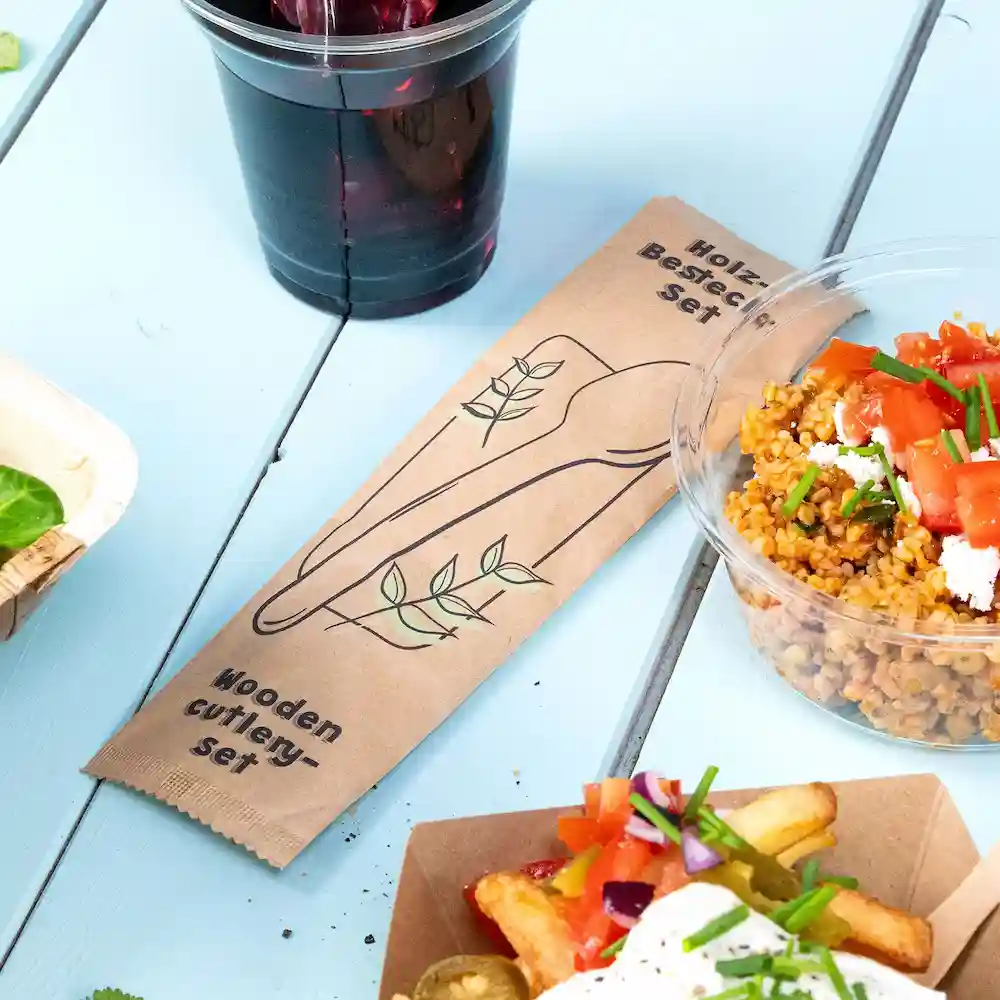 Verpakking van een houten bestekset naast een plastic beker, een kom met graansalade en een kartonnen doos friet op een blauwe ondergrond.