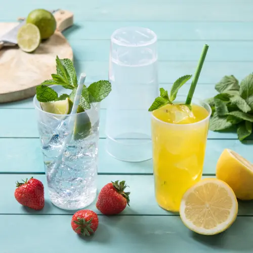 Twee herbruikbare bekers met drankjes, een doorzichtig met een rietje met limoen en munt, en een gele drank met een rietje, omringd door verse aardbeien en citroenschijven.
