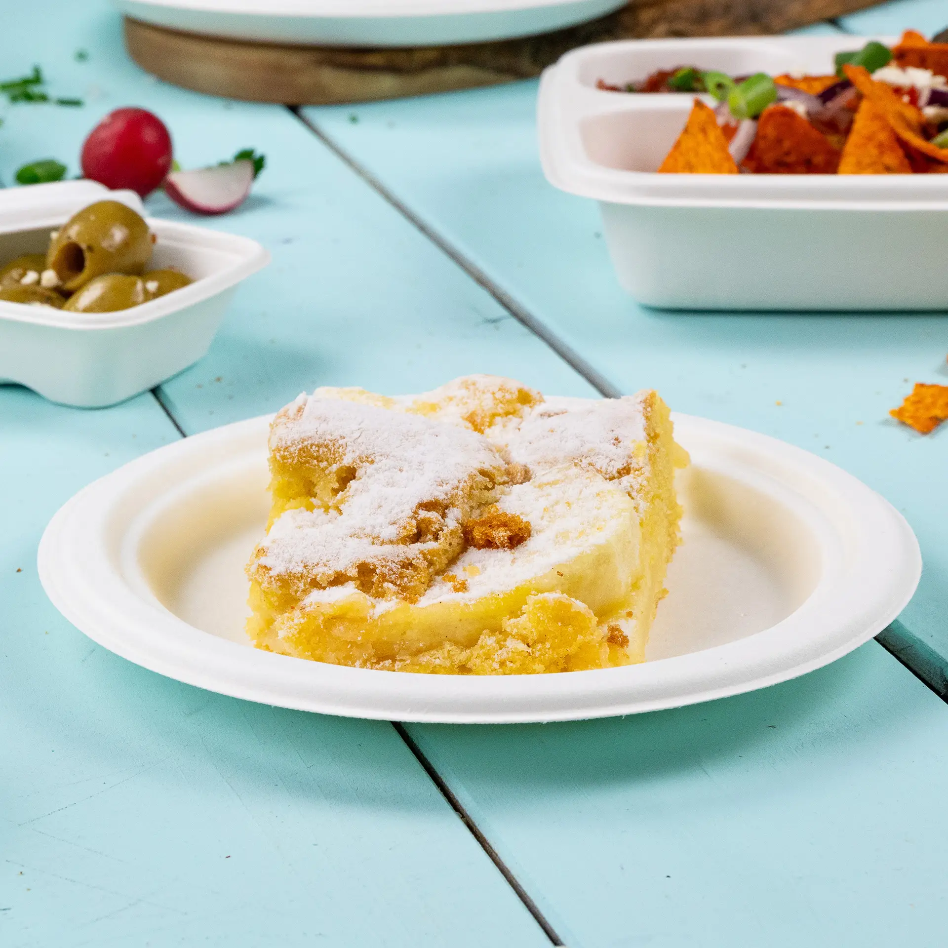 Een plak cake bestrooid met poedersuiker op een rond biologisch afbreekbaar suikerborden tegen een blauwe achtergrond.