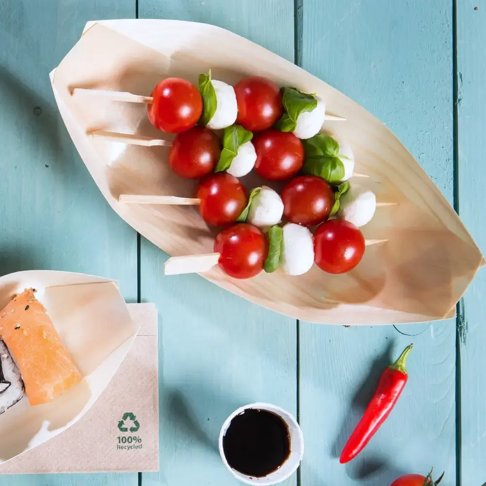 Houten bootvormige schaal met spiesjes van cherrytomaten, mozzarella-balletjes en basilicumblaadjes, naast sushi en dipsaus.