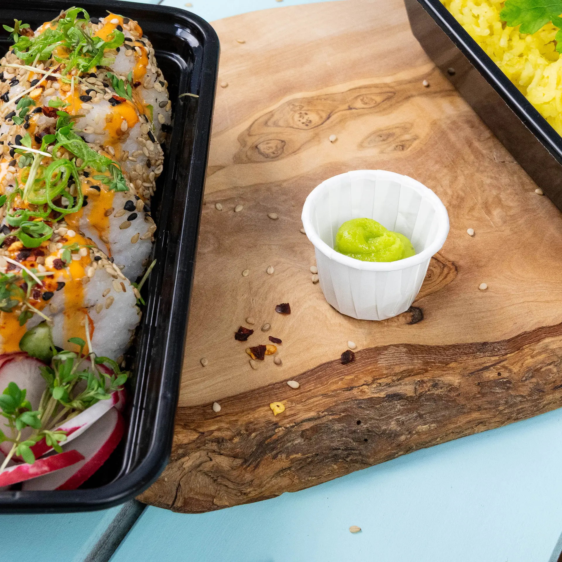 Container met sushirollen met sesamzaad en toppings, vergezeld van een klein wit papieren bekertje met wasabi.