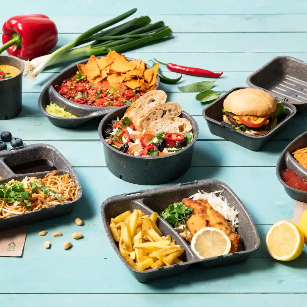 Diverse voedingsmiddelen in zwarte containers: nacho's, salade met brood, spaghetti, frietjes, een hamburger en extra ingrediënten op een houten tafel.