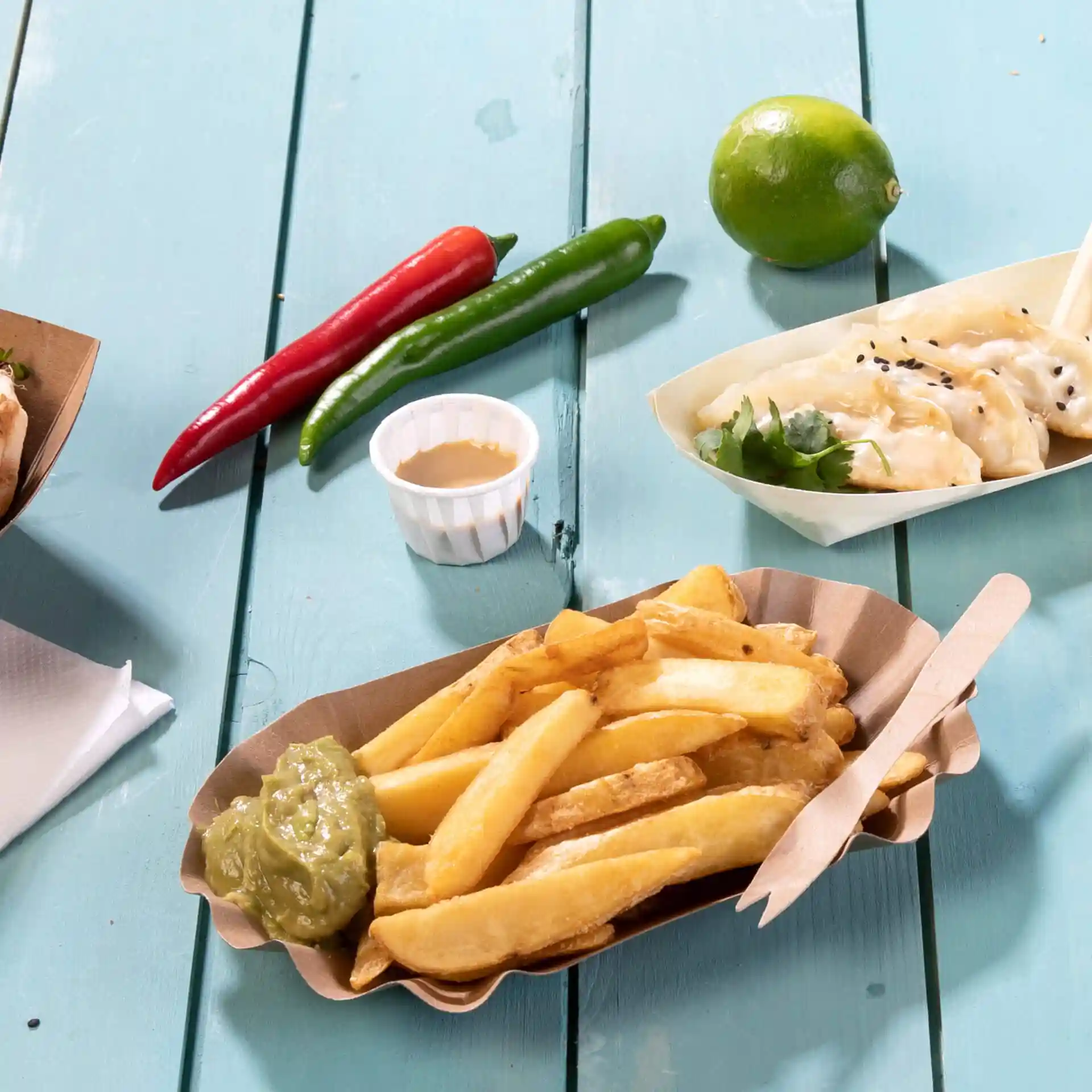 Afbeelding van een portie friet met groene saus in een papieren bakje, omringd door chilipepers, een limoen en een klein bakje saus.