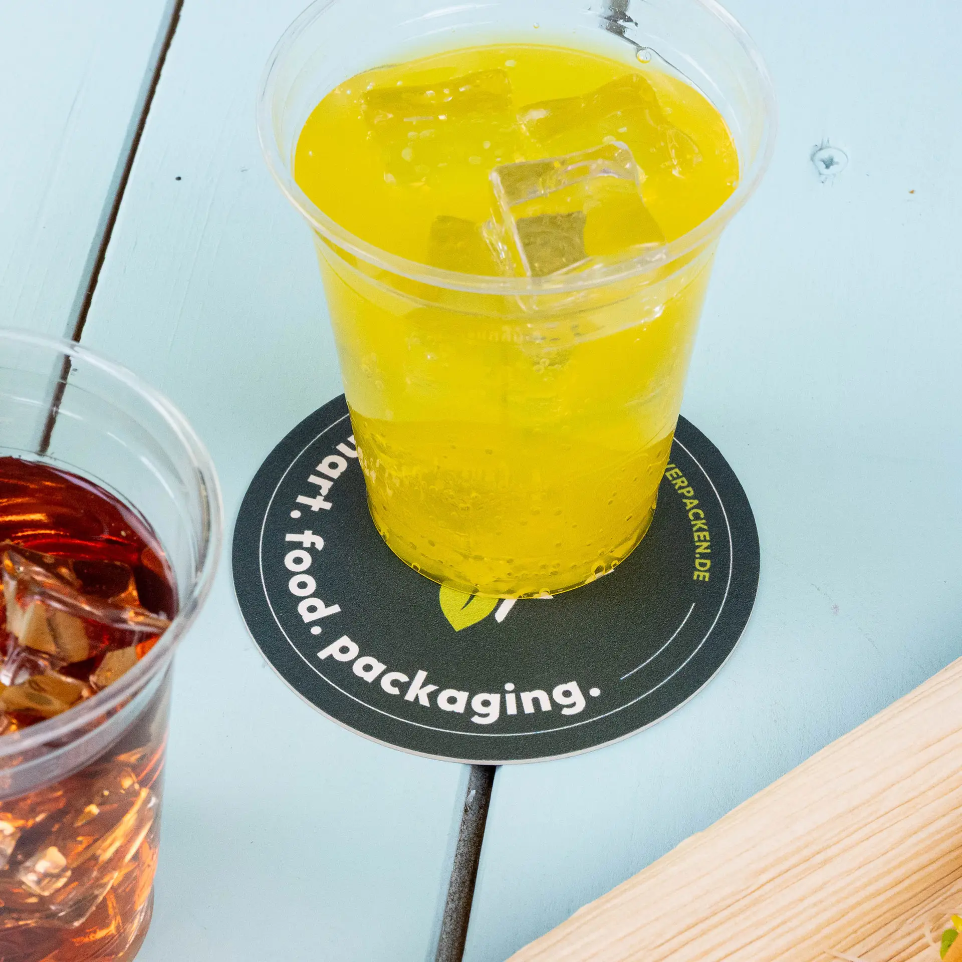 Geel drankje in een heldere beker op een ronde bedrukte onderzetter met tekst, naast een beker met een donker drankje met ijs.