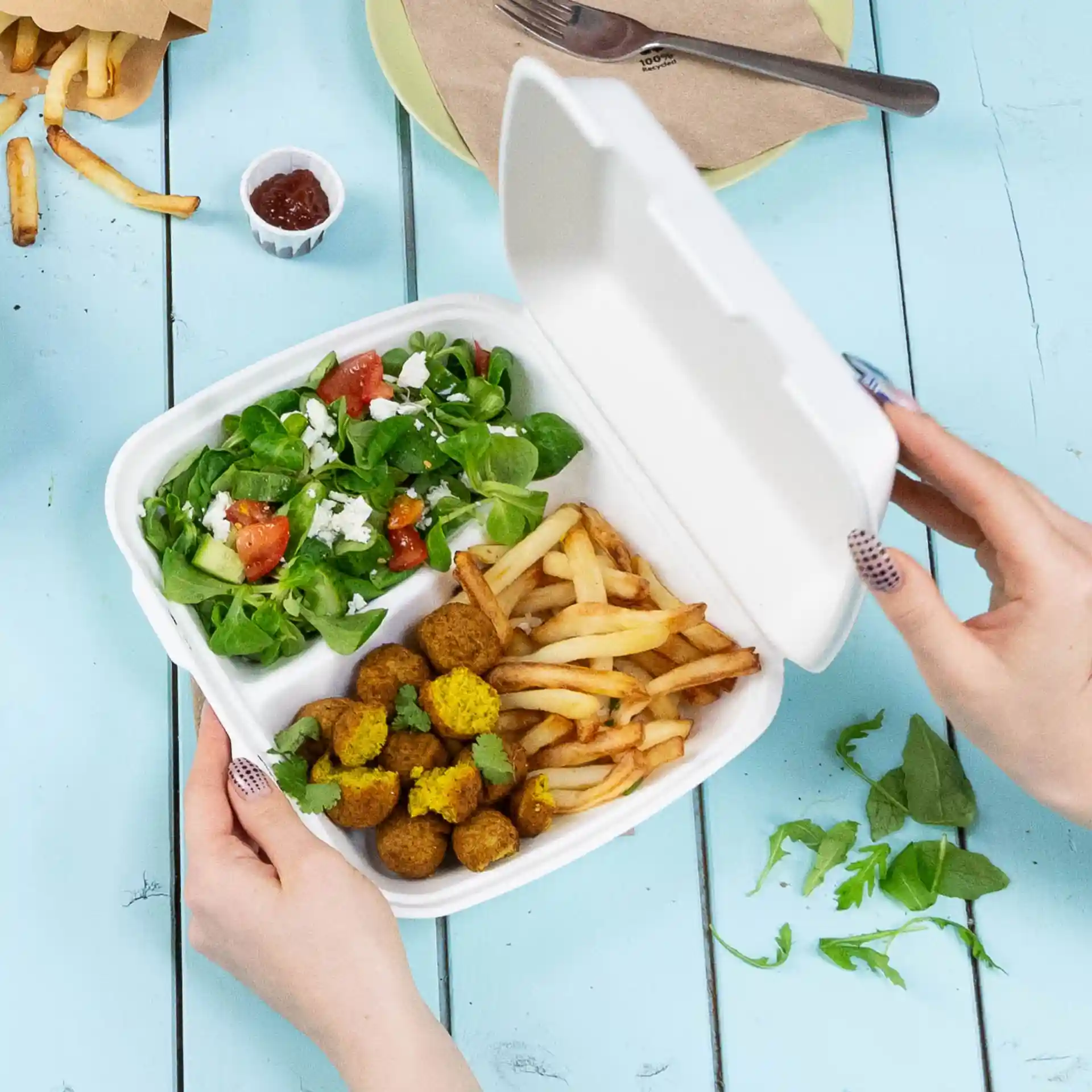 Een hand houdt een witte container met scharnierdeksel met twee compartimenten, gevuld met salade, friet en falafelballen.