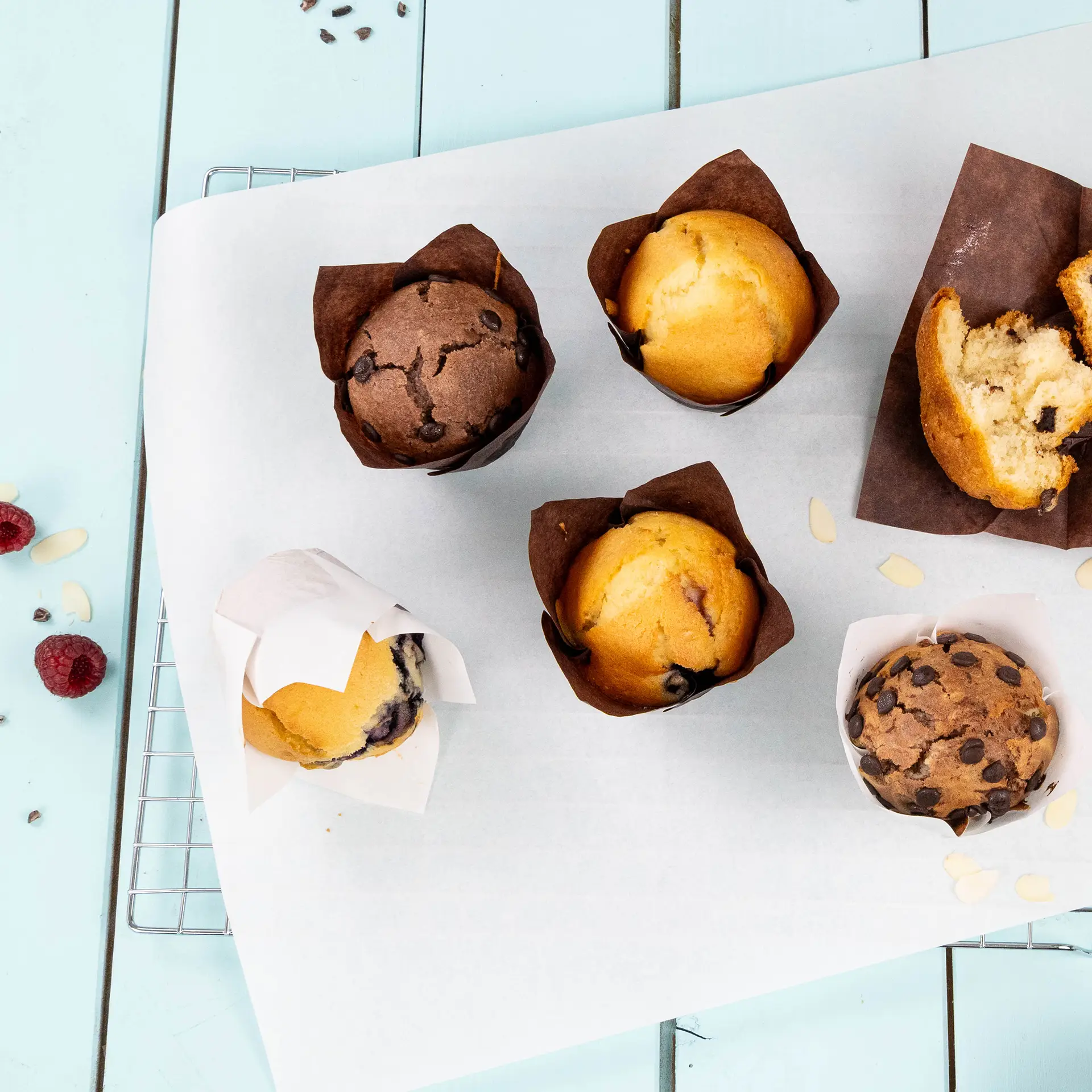 Een chocolade-muffin en vijf vanille-muffins in bruine vetvrije papieren cups op een met bakpapier beklede schaal. Frambozen en geschaafde amandelen erbij.