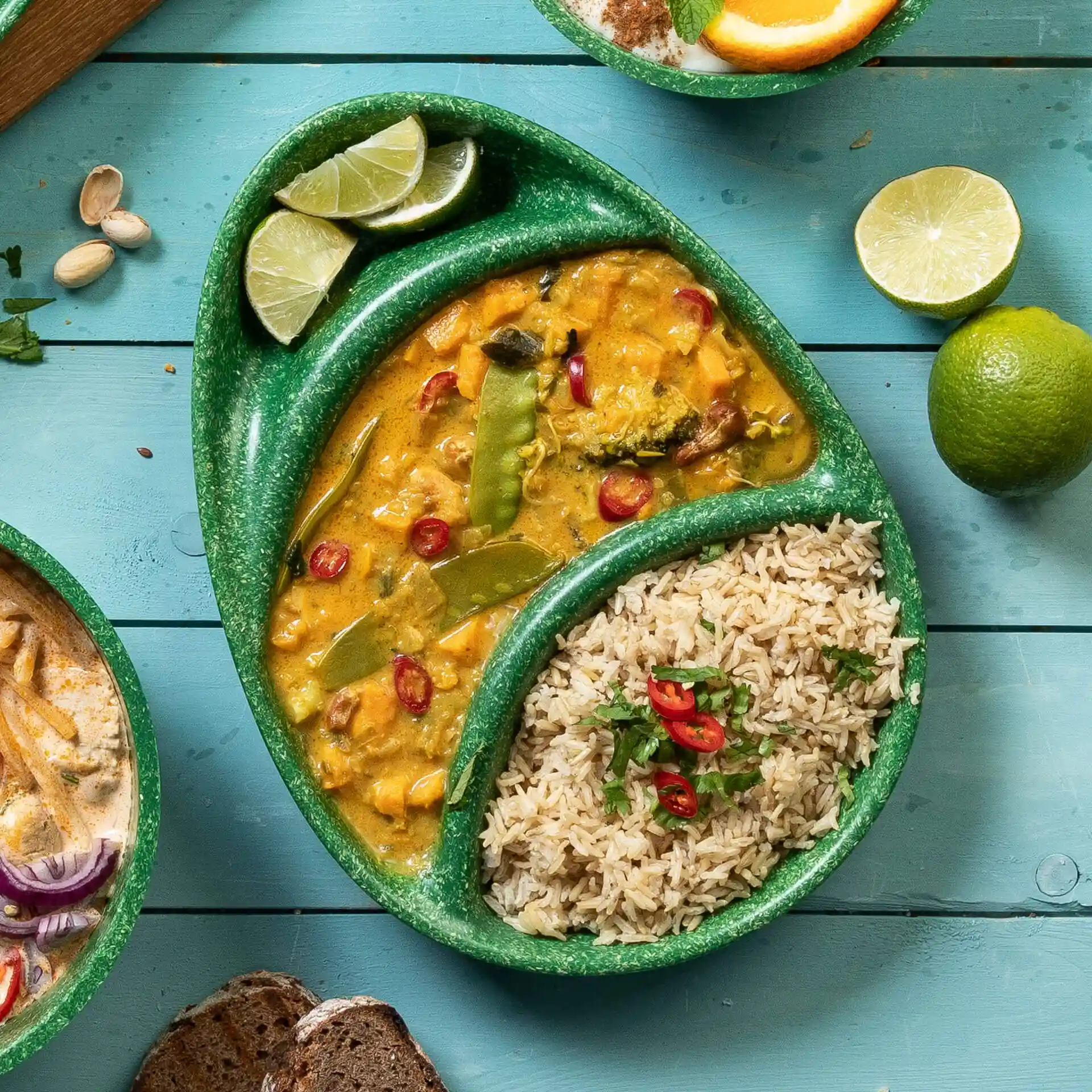 Groen herbruikbaar bord met drie compartimenten, gevuld met curry, rijst en limoenpartjes op een blauwe houten ondergrond.