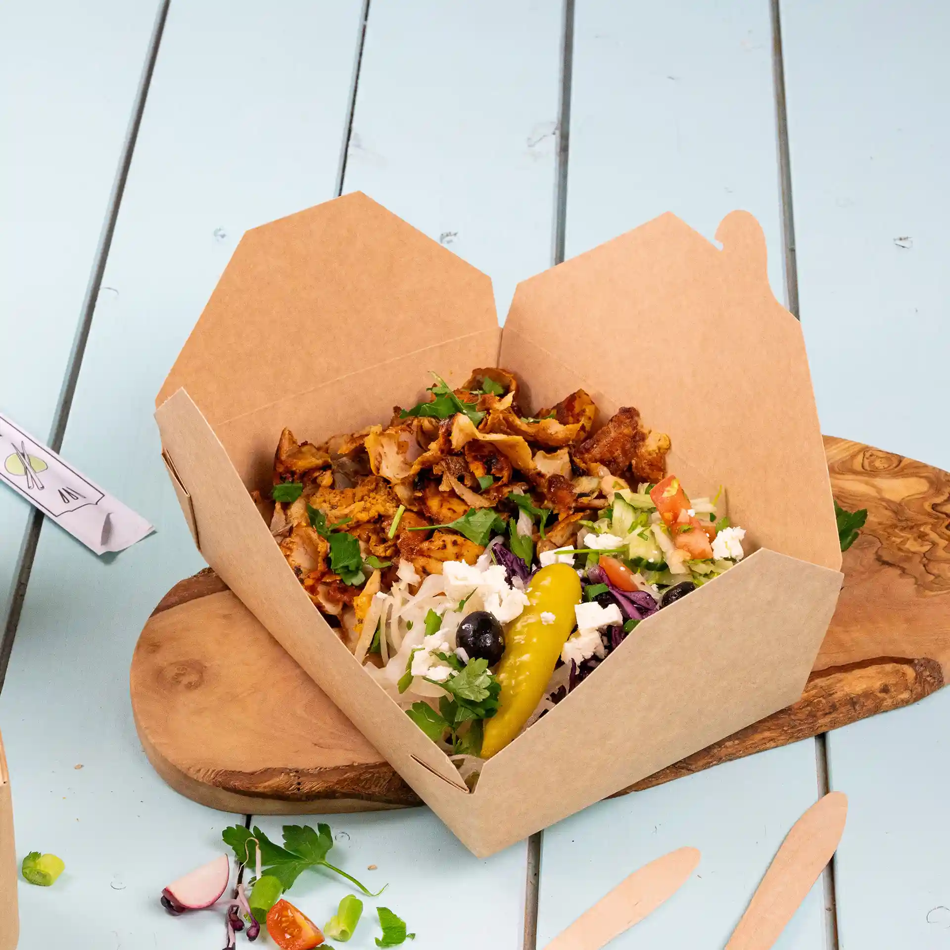 Bruine kartonnen takeaway-container gevuld met eten, waaronder vlees, salade en een augurk, op een houten plank.