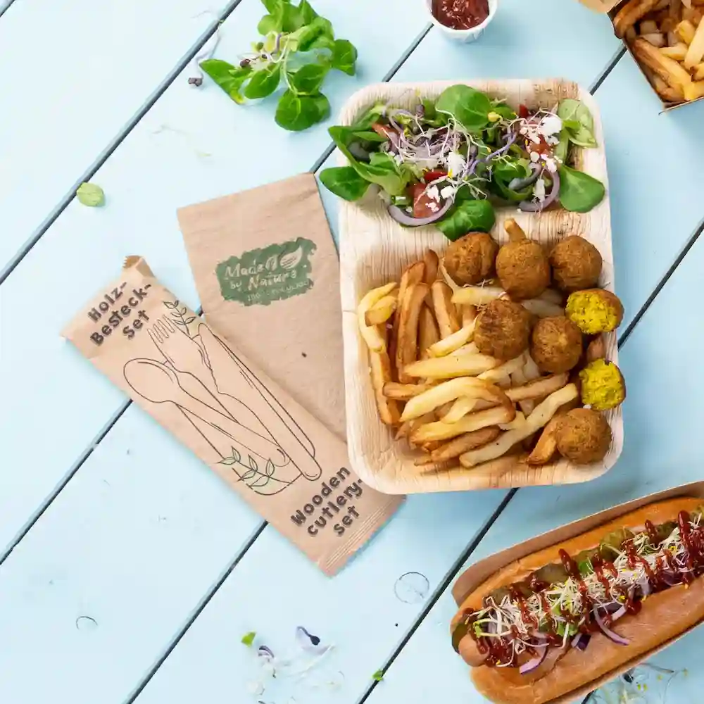 Houten bestekset naast een voedselplaat met salade, frietjes, falafelballen en een hotdog op een blauwe tafel.