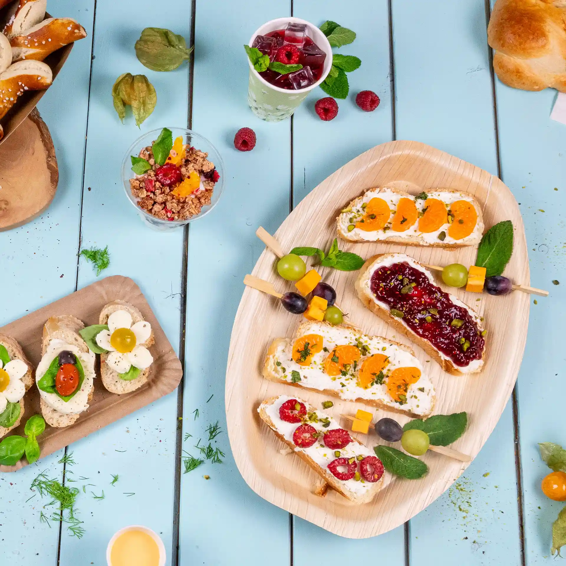 Rechthoekig palmblad dienblad met verschillende open sandwiches, gegarneerd met fruit en kruiden, op een blauwe houten tafel.