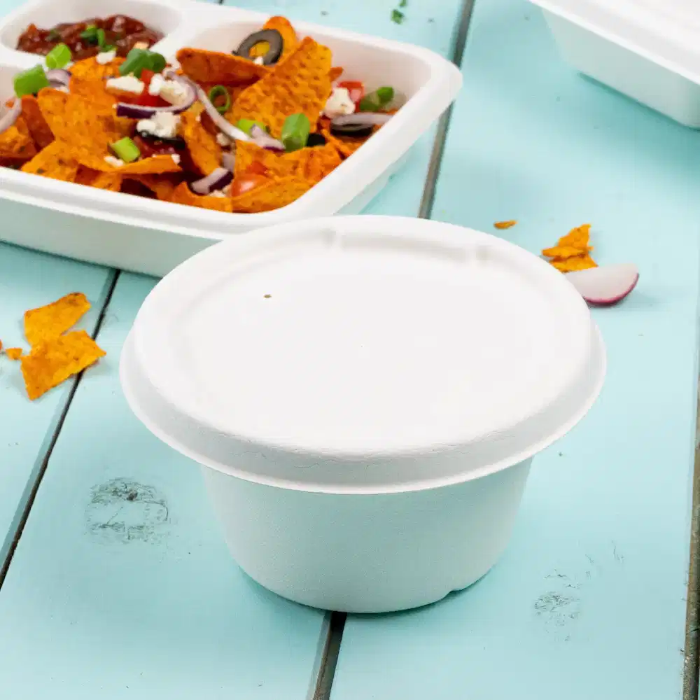 Witte papieren soepbeker op een blauwe tafel met op de achtergrond nacho's en een afgesloten voedselcontainer.