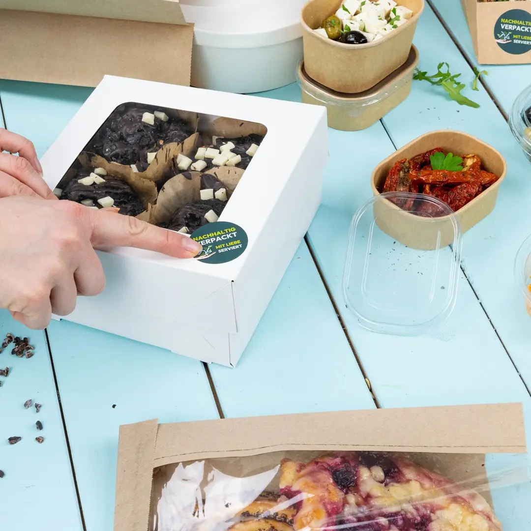 Witte doos met gebakken goederen en een doorzichtig deksel, omringd door verschillende voedselcontainers op een blauwe tafel.