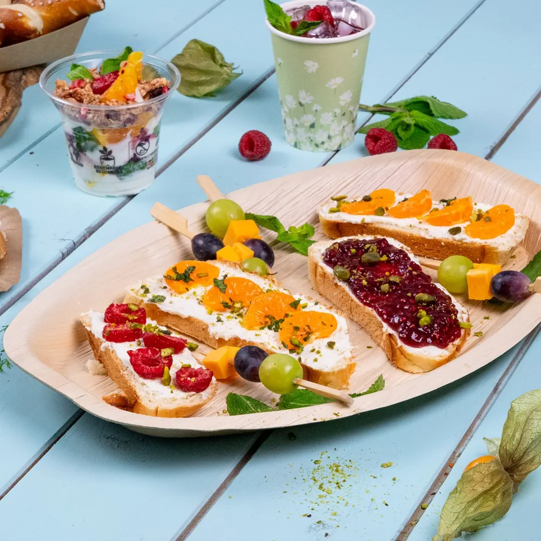 Plank met verschillende fingerfood, waaronder brood met roomkaas, fruit en een fruitspies, naast een yoghurtparfait.