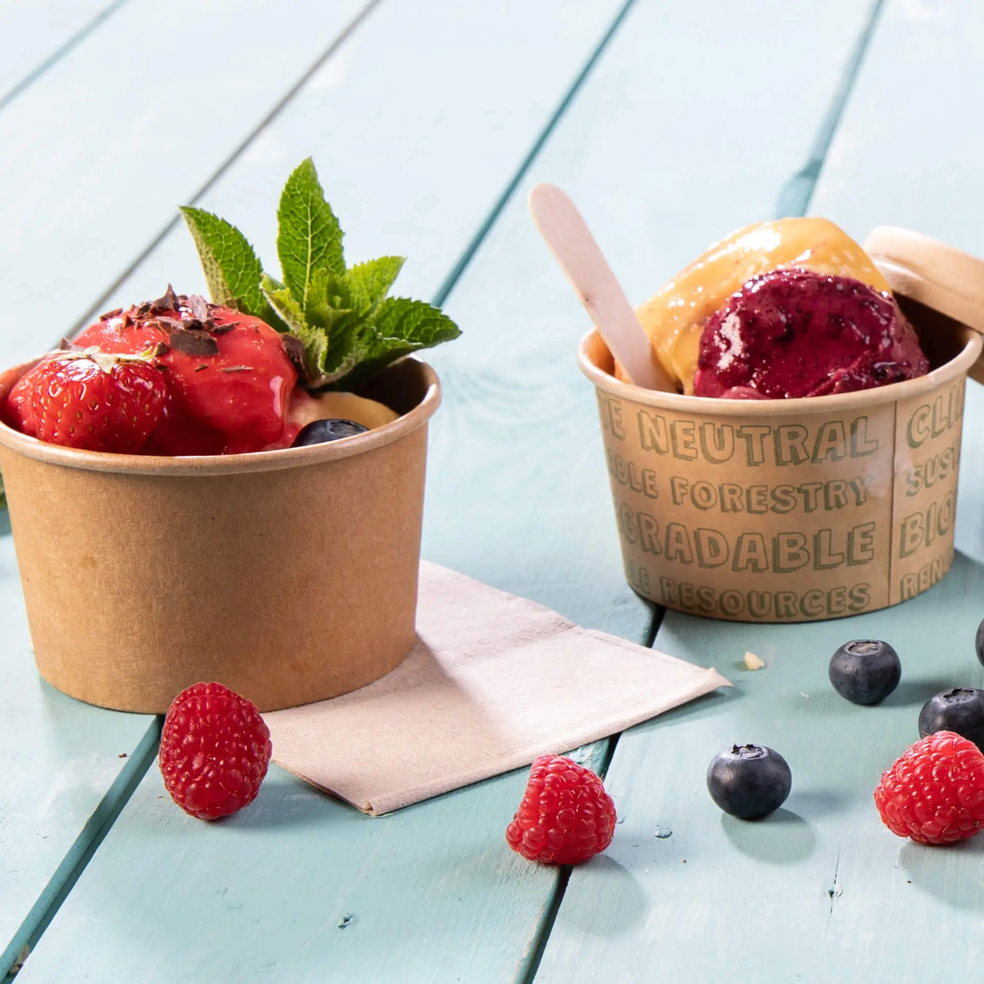 Twee papieren bekers met ijs en fruittoppings, de ene met aardbeien en munt, de andere met paarse en gele ijs.