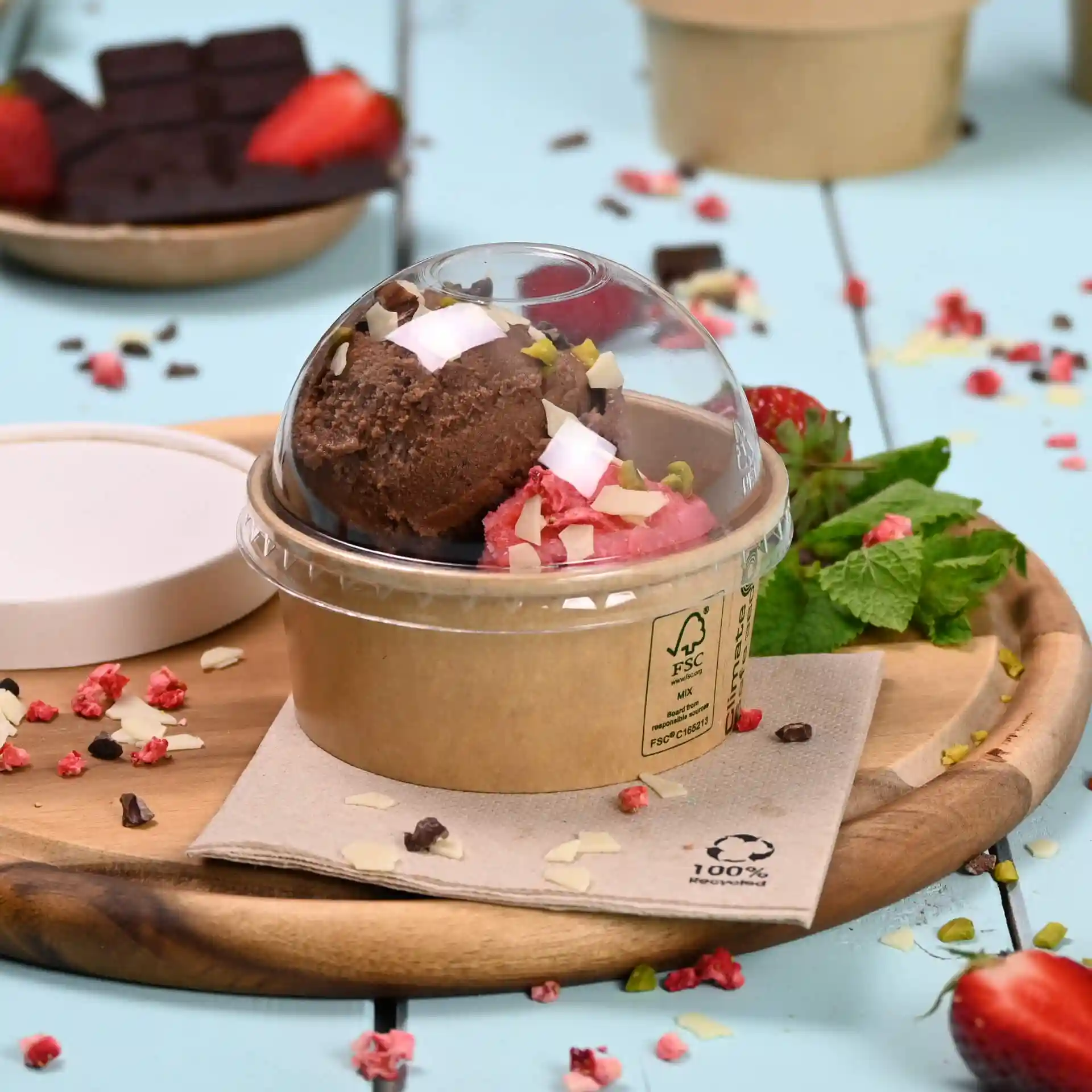 Container met twee bollen ijs, getopt met noten en kokos, op een houten plank met munt en aardbeien.