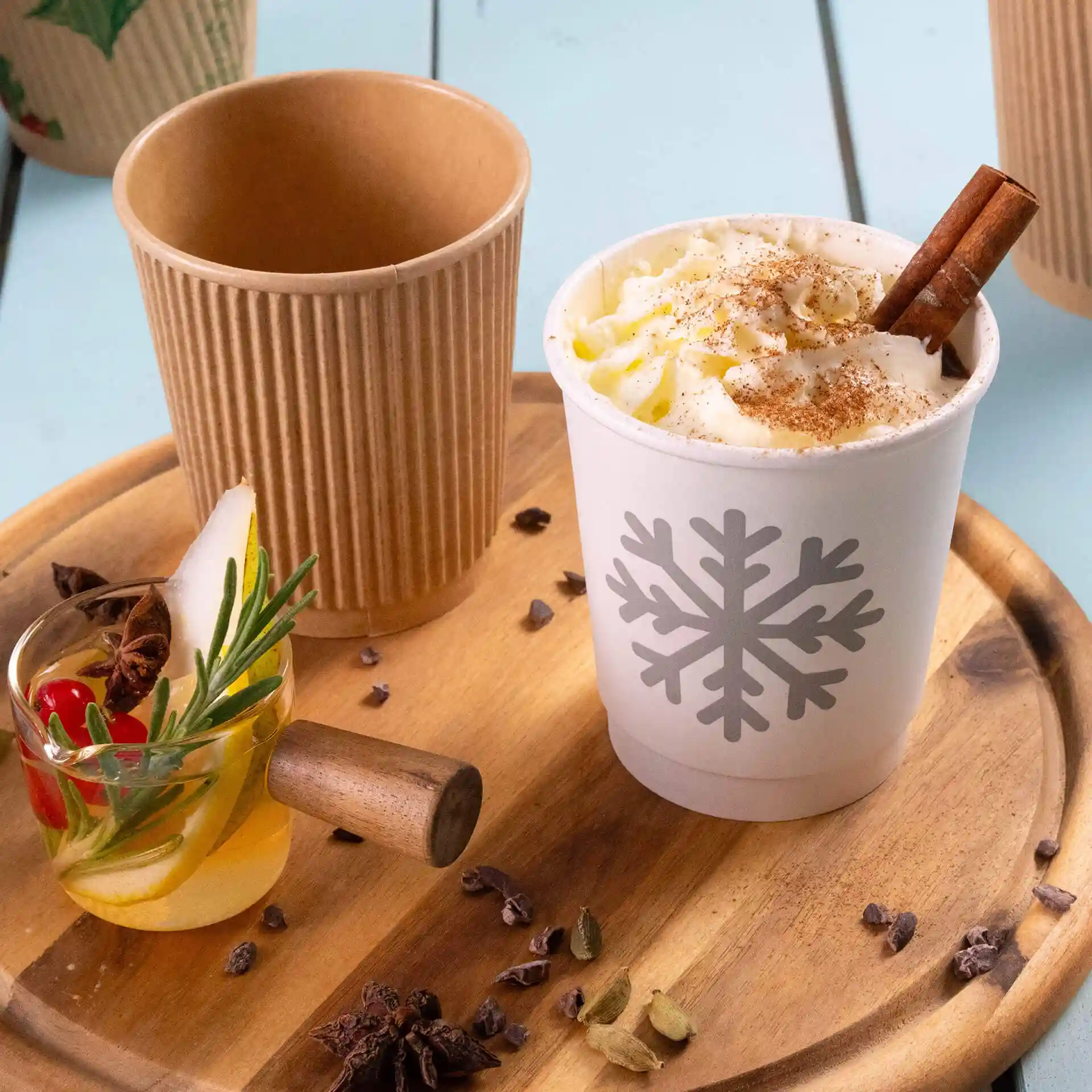 Witte beker met sneeuwvlokdesign, gevuld met slagroom en een kaneelstokje, naast een kraftbeker en een gegarneerde drank.