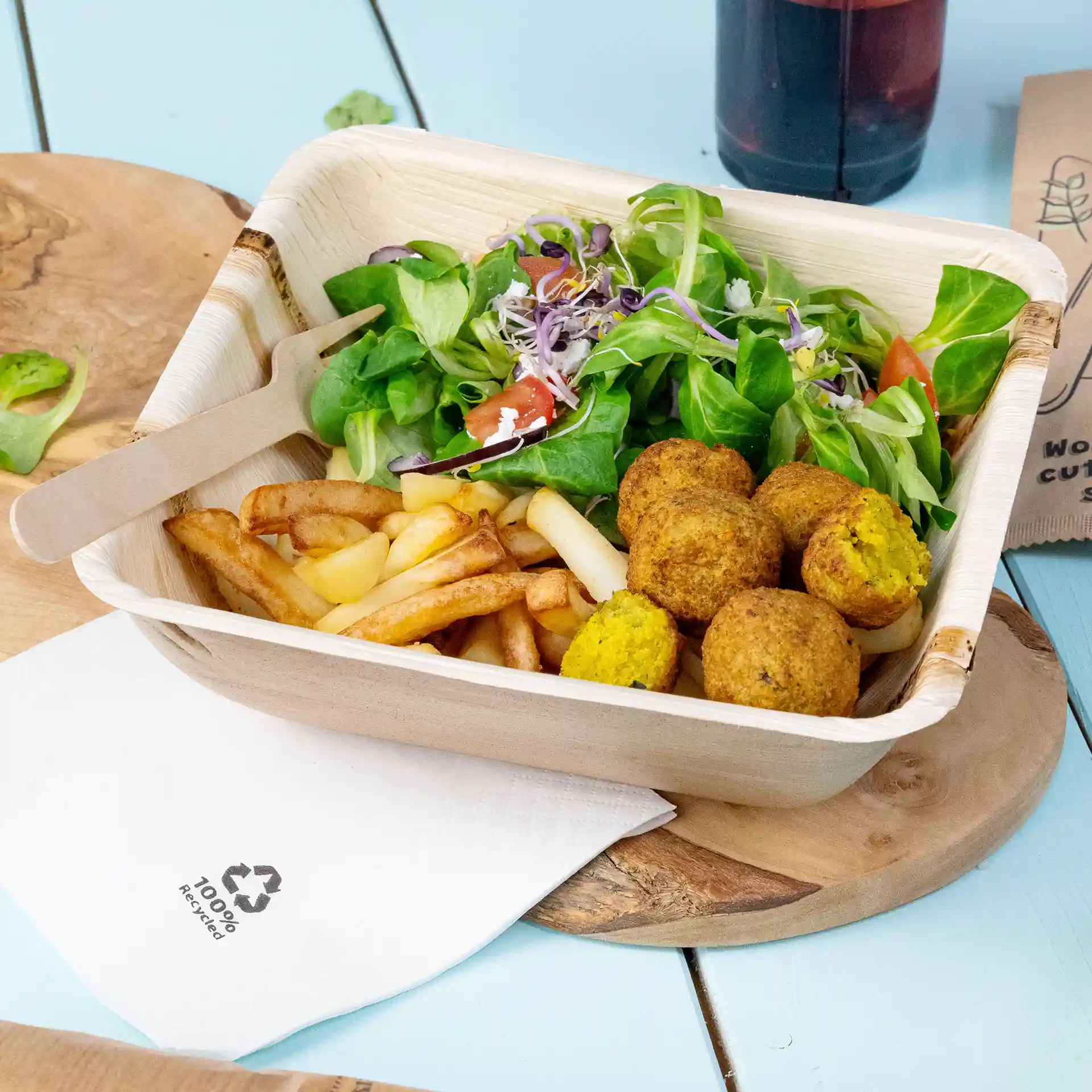 Diepe vierkante palmbladkom met salade, friet en falafelballen, met een houten vork en een servet ernaast.