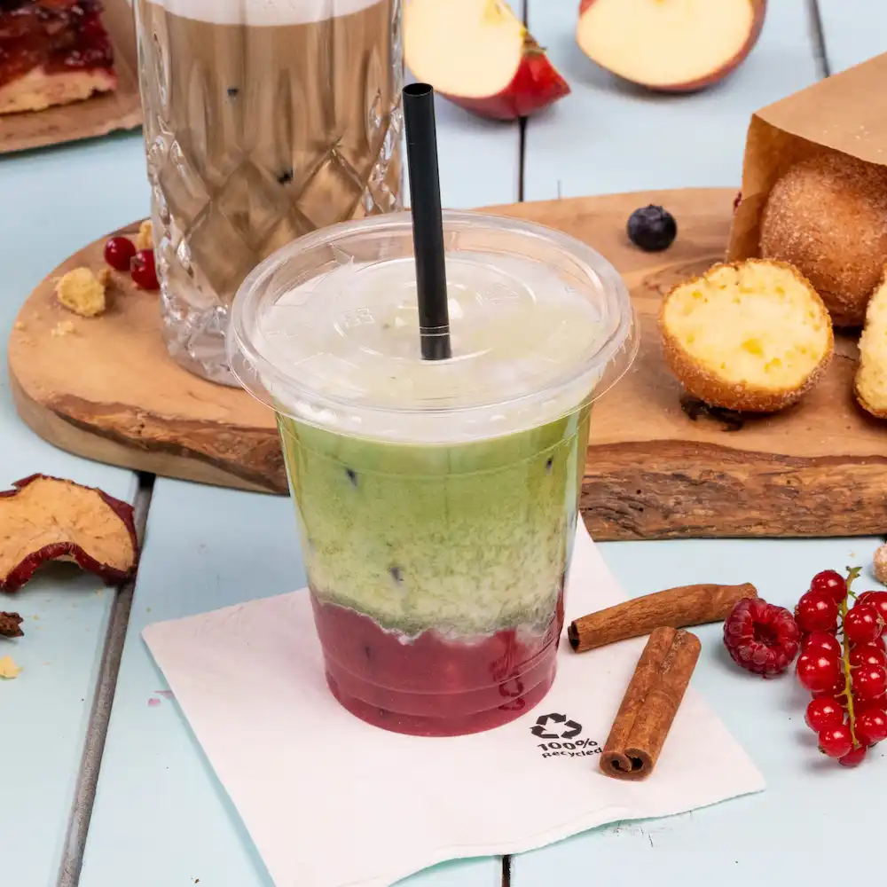 Gelaagde ijsdrank in een doorzichtige beker met een rietje, omringd door snacks en een servetje op een lichtblauwe ondergrond.