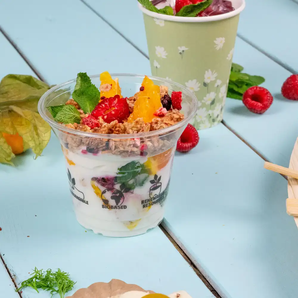 Heldere plastic dessertbeker gevuld met yoghurt, fruit en granola, op een blauwe ondergrond met muntbladeren erbij.