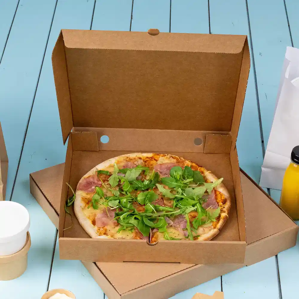 Pizza in een kartonnen afhaaldoos, belegd met ham en gemengde groenten op een blauwe houten ondergrond.