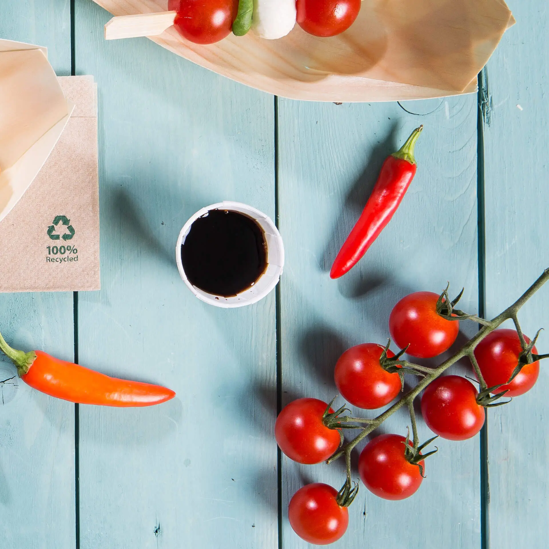 Afbeelding van een houten bord met cherrytomaten, chilipepers, een klein wit papieren bekertje met donkere saus en een bruine verpakking met recycling-symbool.