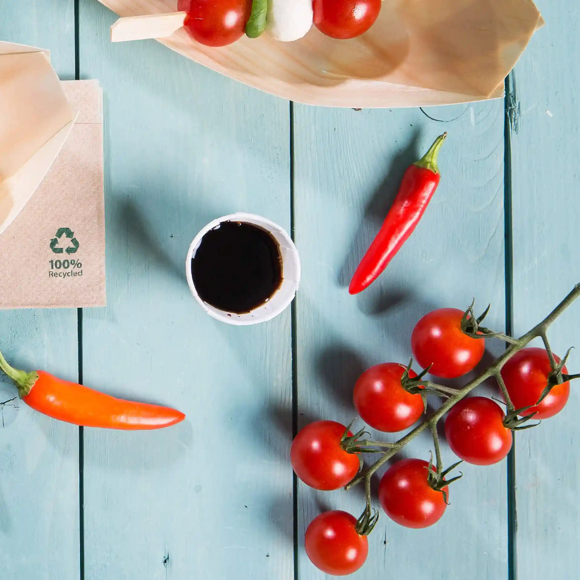 Afbeelding van een papieren beker met donkere vloeistof, omringd door cherrytomaten, chilipepers en een houten bord op een blauwe houten ondergrond.