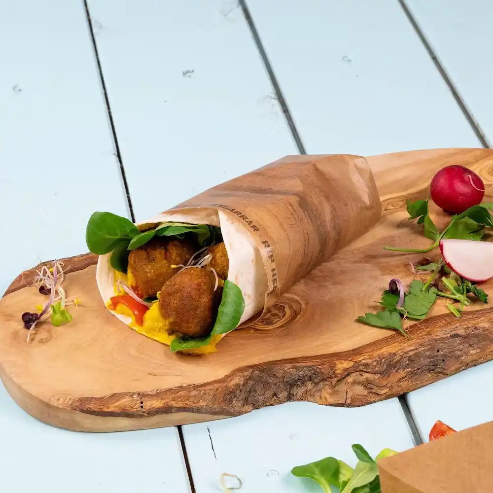Een gewikkelde snack gevuld met falafel, spinazie en saus op een houten plank, vergezeld van radijzen en kruiden.