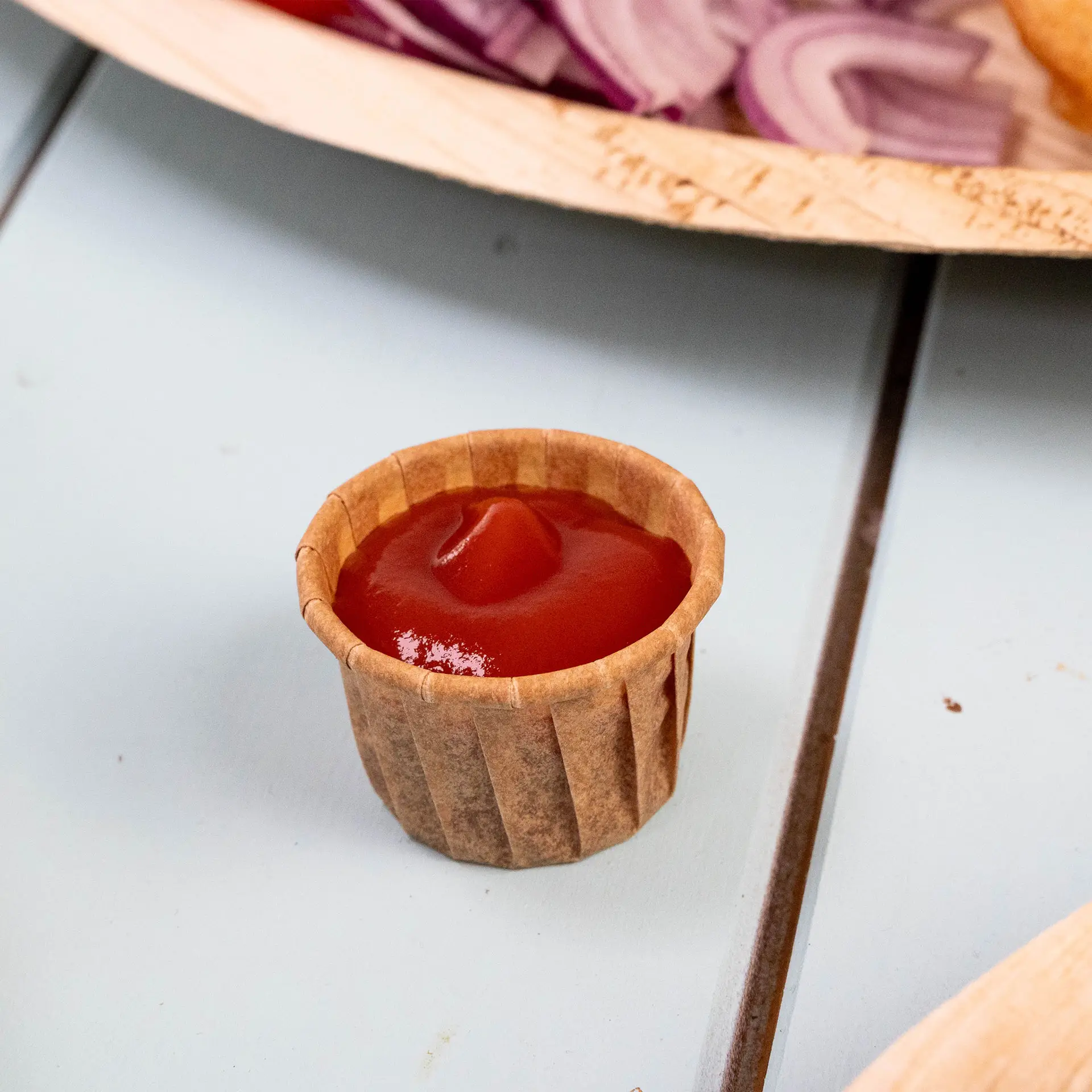 Bruine papieren beker van 15 ml gevuld met ketchup, op een blauwe ondergrond, met gesneden uien en een bord op de achtergrond.