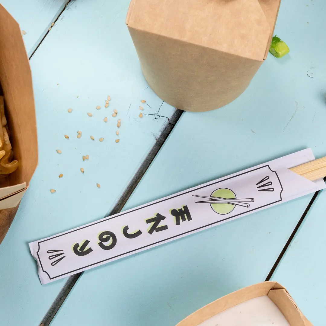 Chopstick-verpakking met grafisch ontwerp en chopsticks op een blauwe houten ondergrond, omringd door voedselcontainers en sesamzaad.