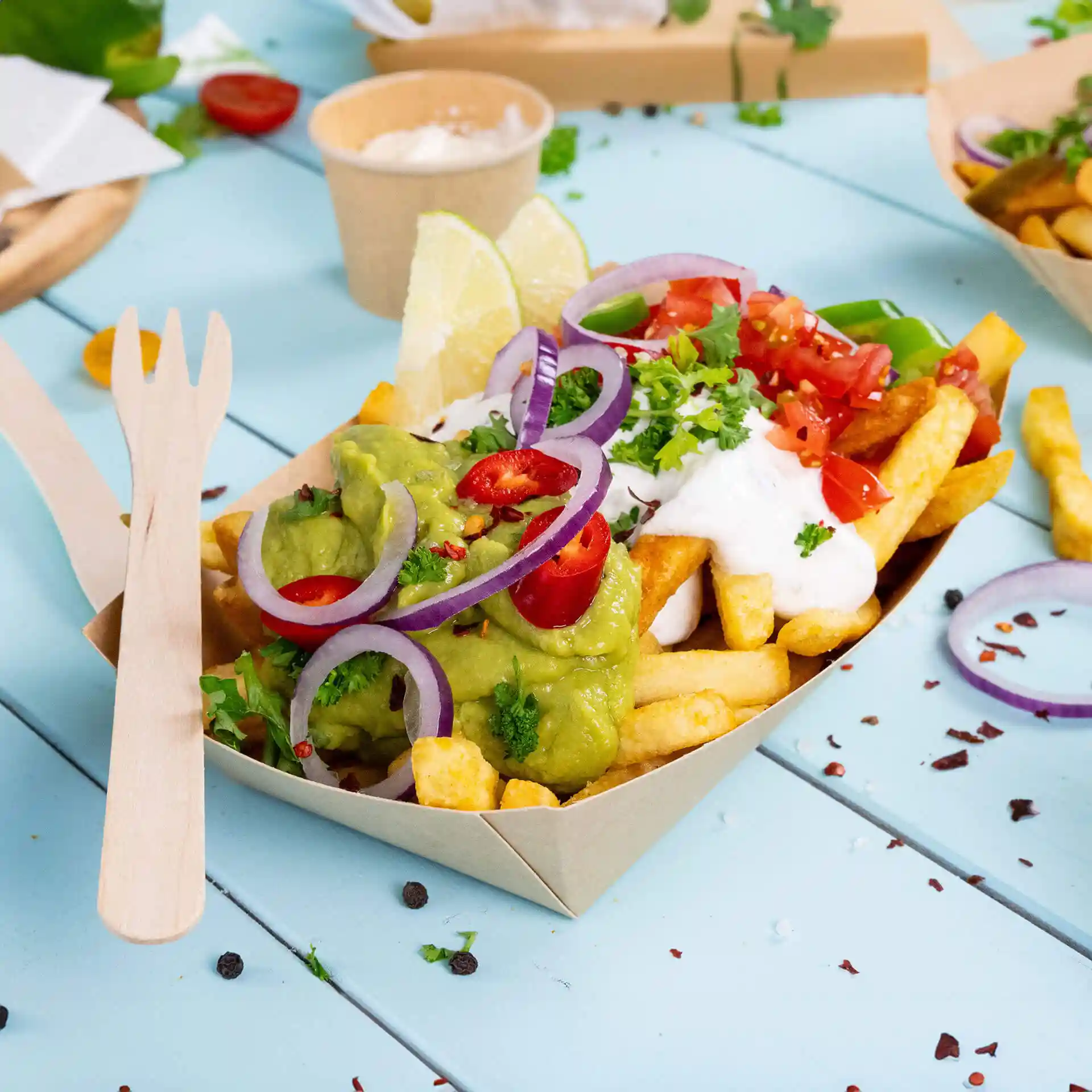 Diepe kartonnen snackkom gevuld met frietjes, guacamole, zure room, in blokjes gesneden tomaten en gegarneerd met uien en limoen.