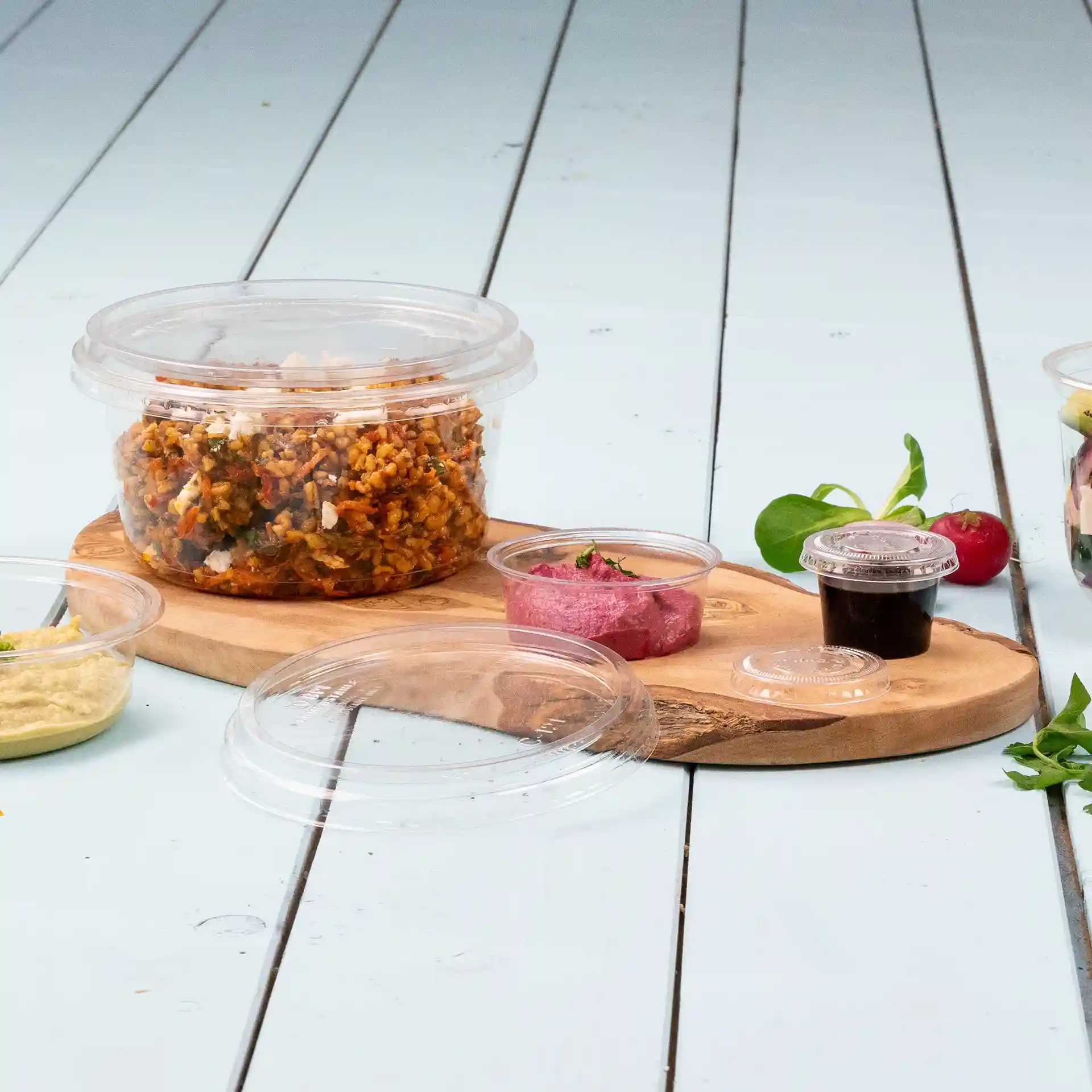 Transparante ronde deli-container met deksel, gevuld met voedsel, op een houten plank met kleine sausbekers erbij.