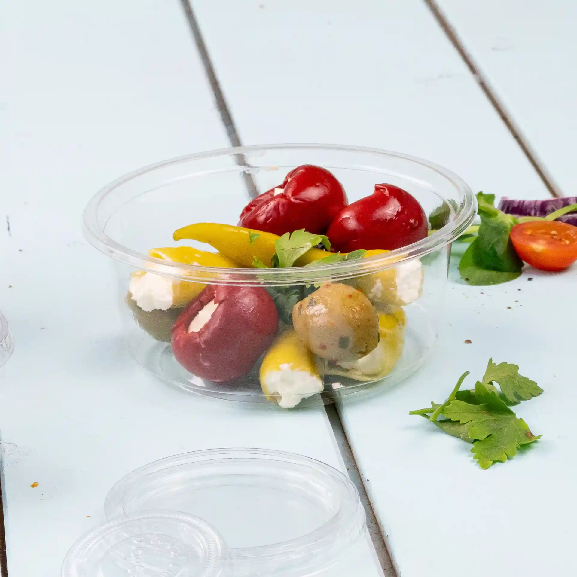 Transparante ronde deli-pot met verschillende paprika's, olijven en kaas, geplaatst op een lichtblauwe houten ondergrond.