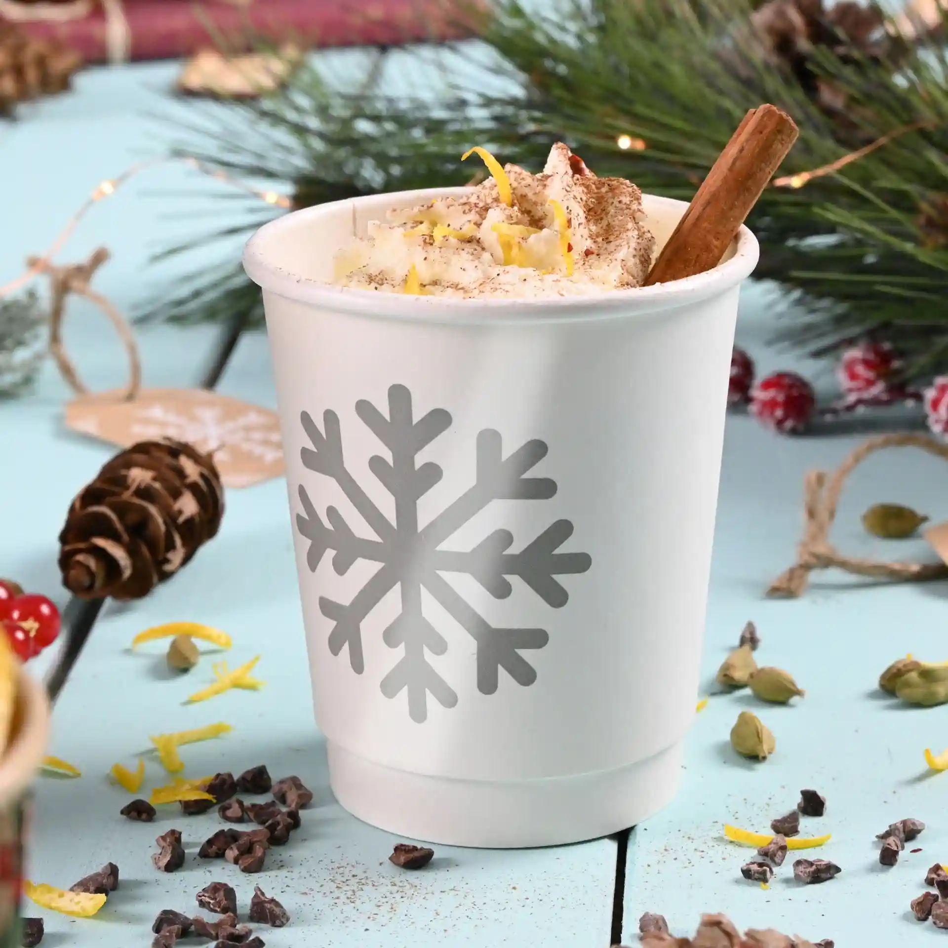 Witte dubbelwandige papieren beker met een grijs sneeuwvlokontwerp, gevuld met slagroom en gegarneerd met een kaneelstokje.
