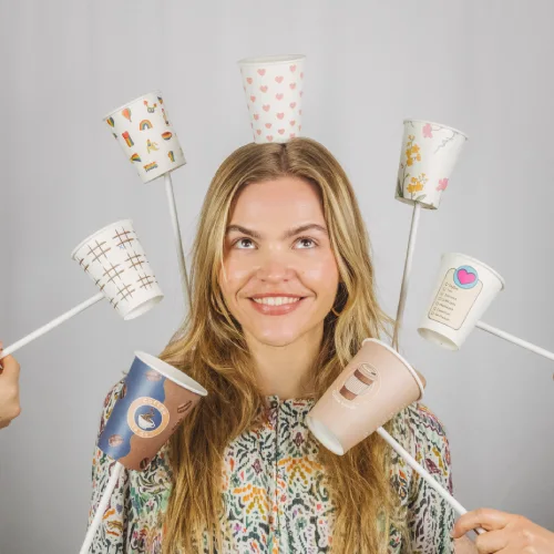 Vrouw met lang haar die glimlacht en verschillende kleurrijke papieren bekers op stokjes rond haar hoofd houdt.
