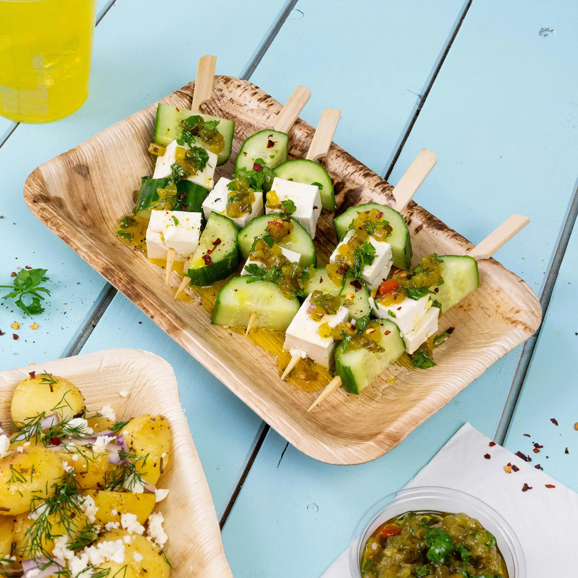 Bamboeschaal met komkommer- en kaasprikkers, gegarneerd met kruiden en saus, geserveerd met aardappelsalade.