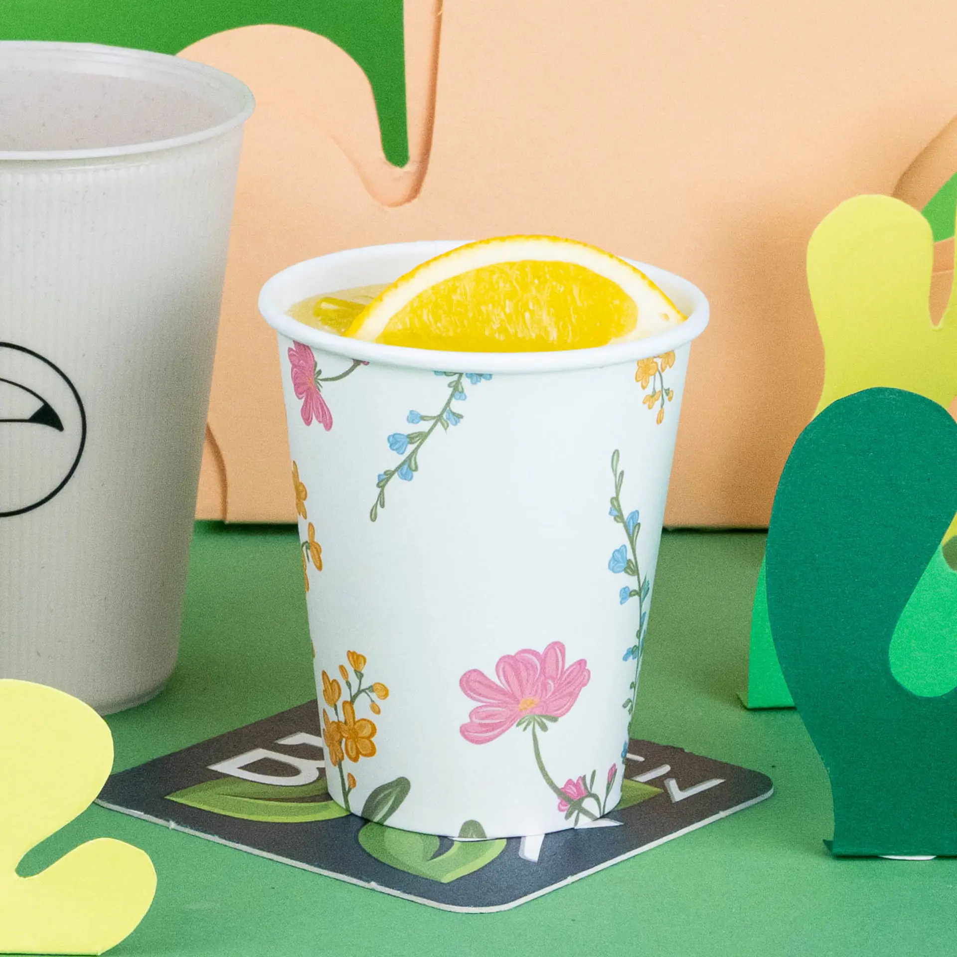 Witte beker met bloemmotief, gevuld met een drankje en een schijfje sinaasappel, op een groene achtergrond met decoratieve elementen.