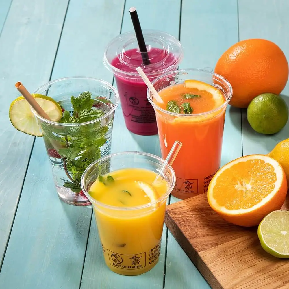 Vier transparante plastic bekers gevuld met kleurrijke dranken, waaronder sinaasappelsap en een roze smoothie, naast verse vruchten.