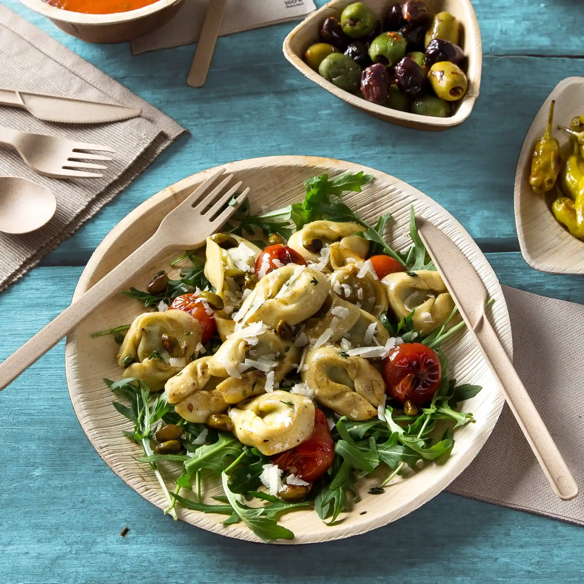 Ronde palmbladplaat met tortellini-salade, rucola en cherrytomaten, vergezeld van houten bestek op een blauwe ondergrond.
