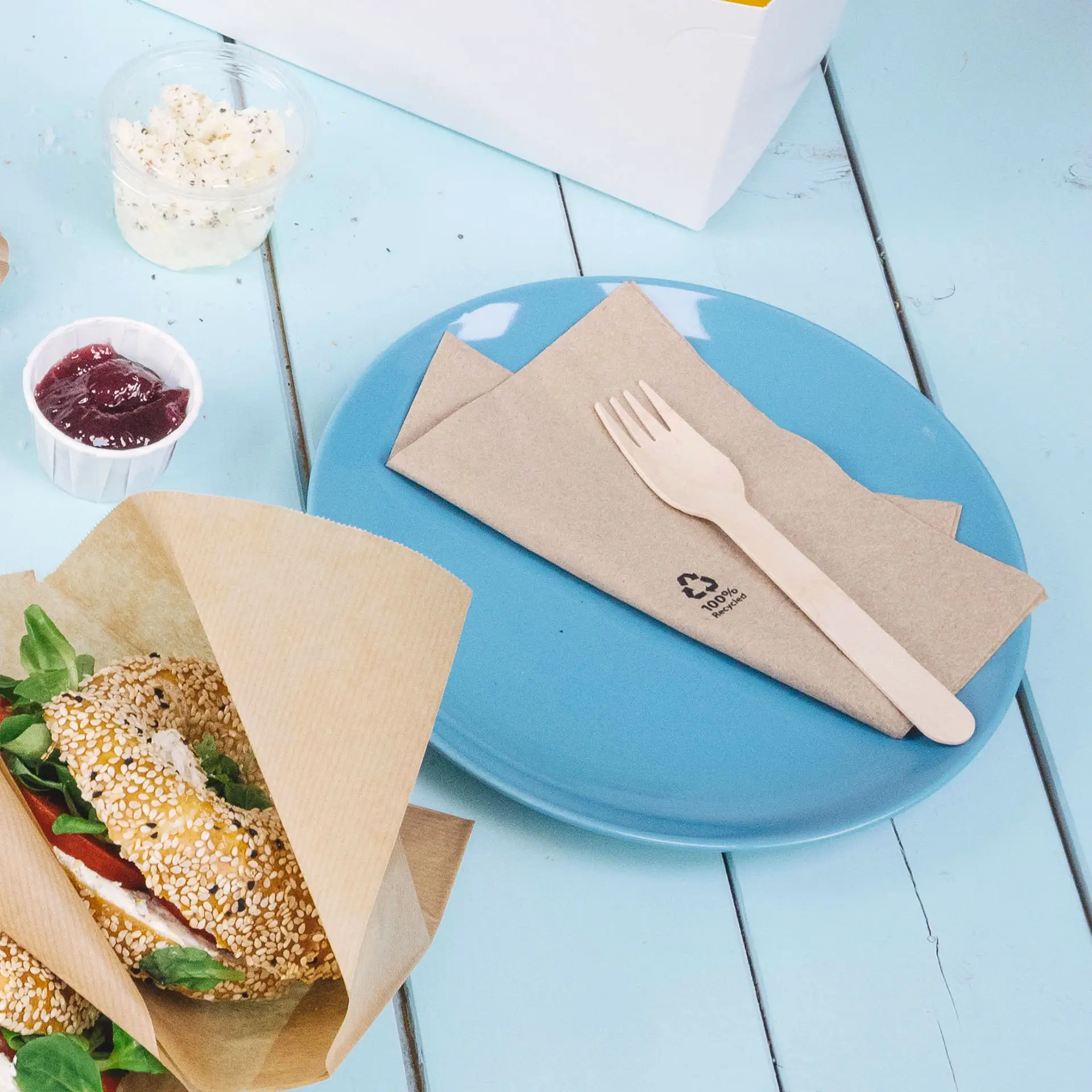 Een blauwe plaat met opgevouwen bruine servetten en een houten vork; een sesam bagel met groenten in de buurt, jam in een beker.