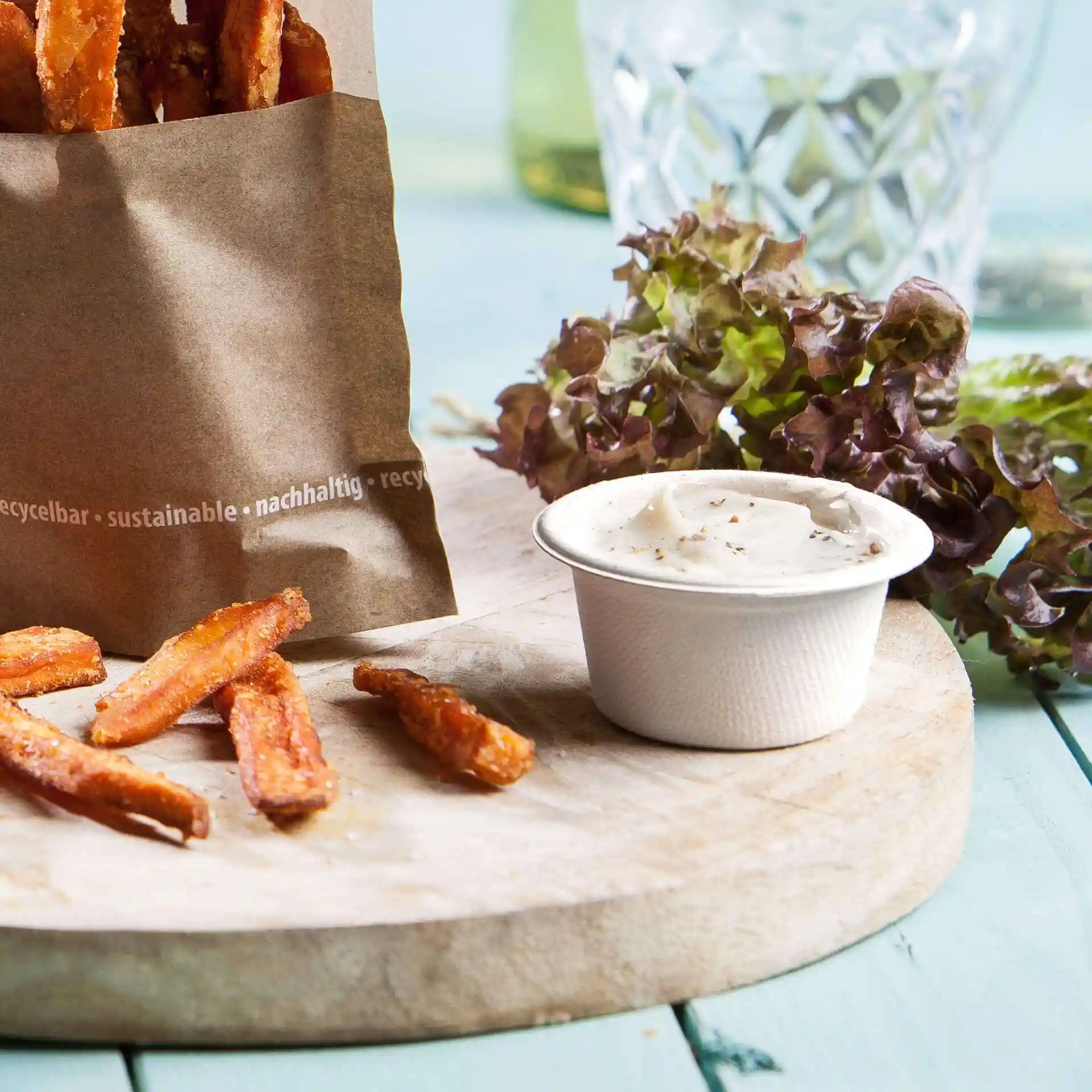 Afbeelding van een papieren zak gevuld met zoete aardappelfrites, een witte dipschaal en bladgroenten op een houten plank.